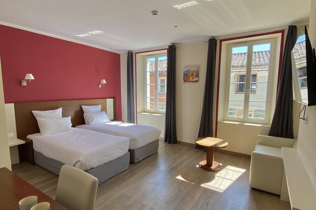 A hotel room with two beds, a small table, and a chair. There are windows with curtains and a view of buildings outside.