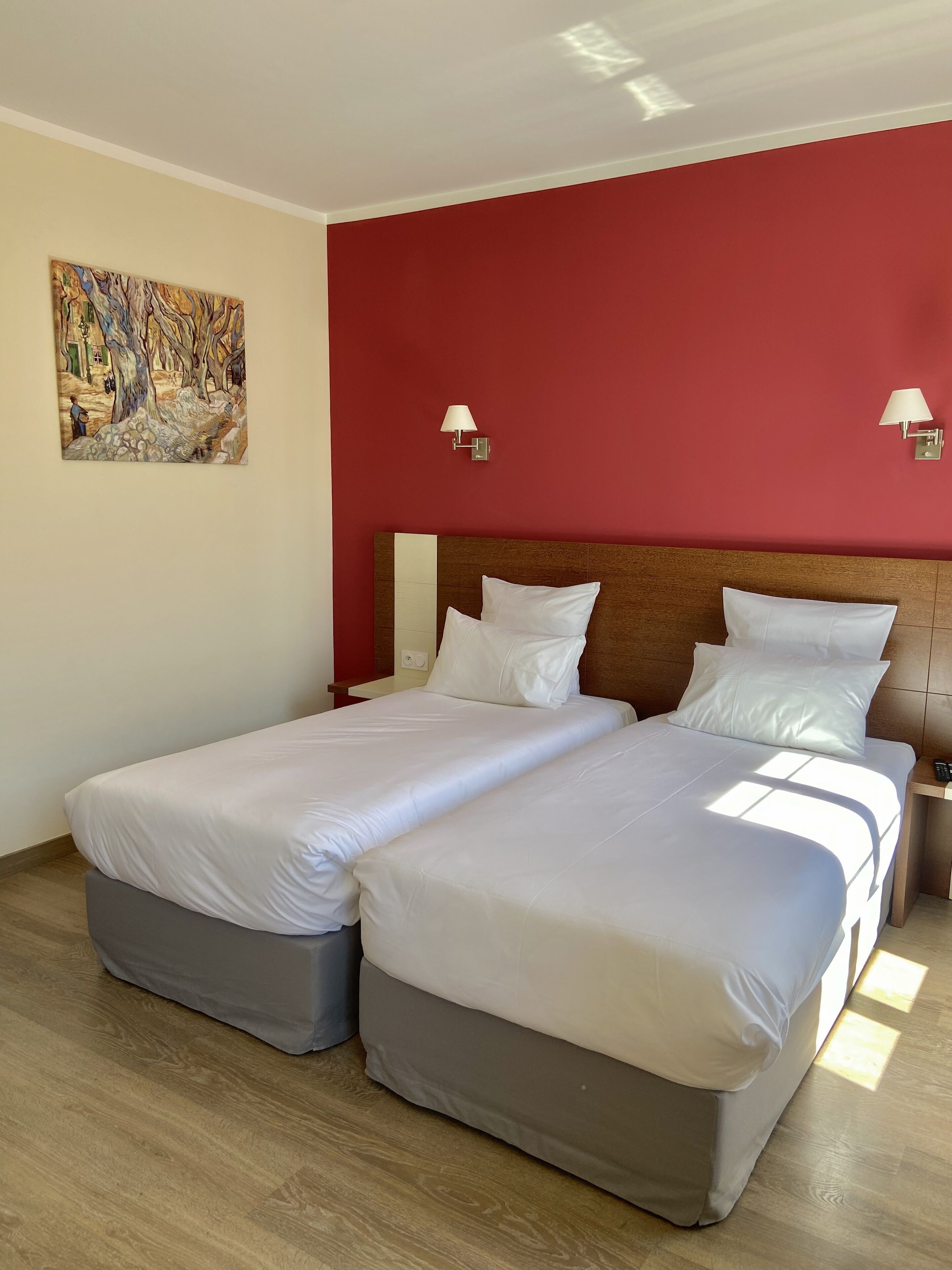 A double bed with white linens in a room with red walls, two wall-mounted lamps, and a painting on the wall.