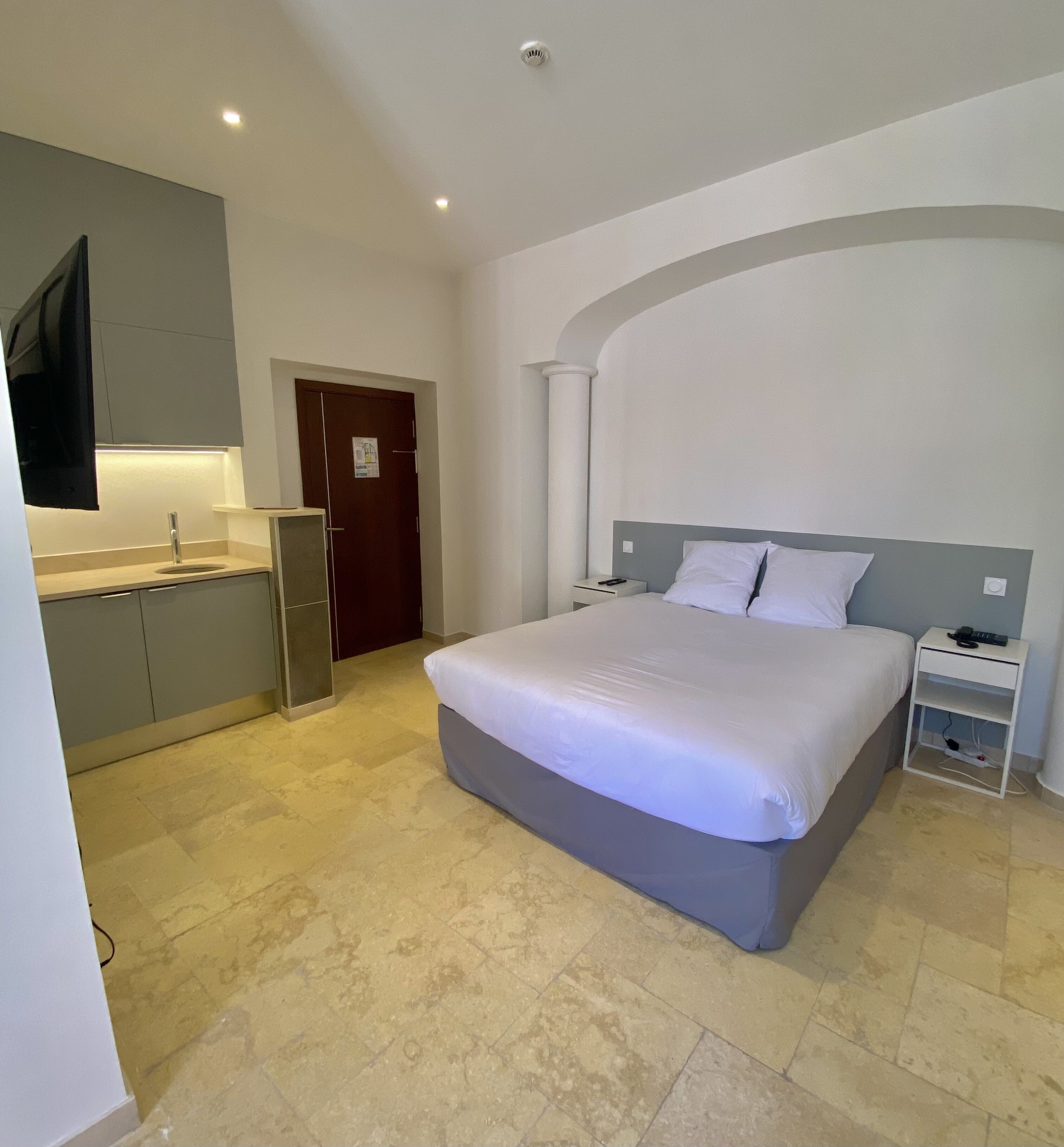 A bedroom with a large bed, a nightstand, a TV mounted on the wall, and a door leading to a bathroom.