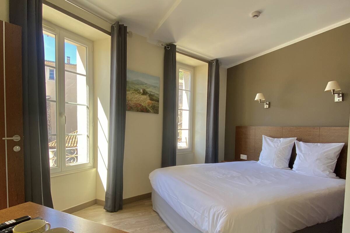 A hotel room with a bed, a desk with two mugs, a painting on the wall, and a window with curtains.