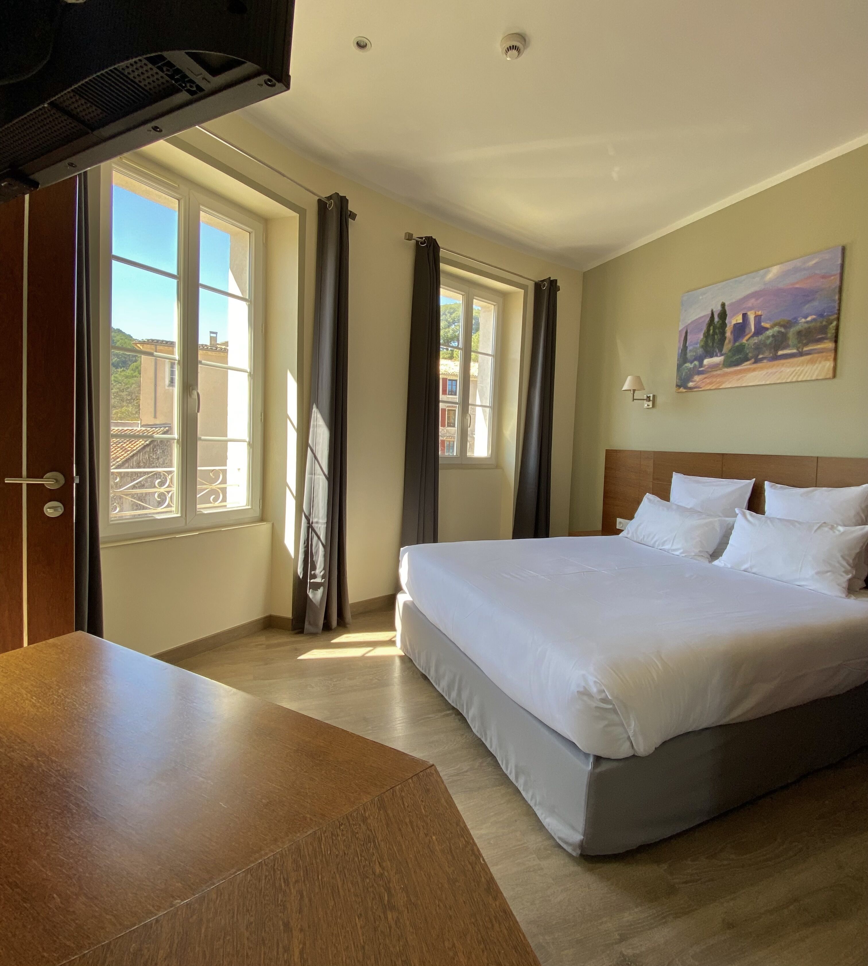 A hotel room with a large bed, a wooden desk, a painting on the wall, and a view of buildings through the window.