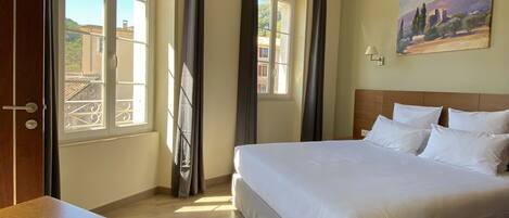 A hotel room with a large bed, a wooden desk, a painting on the wall, and a view of buildings through the window.