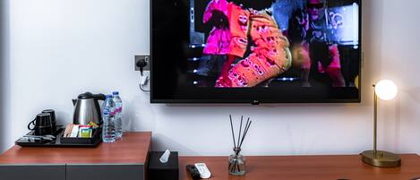 A hotel room with a flat-screen TV mounted on the wall, a wooden desk with a tray of amenities, a remote control, and a lamp.