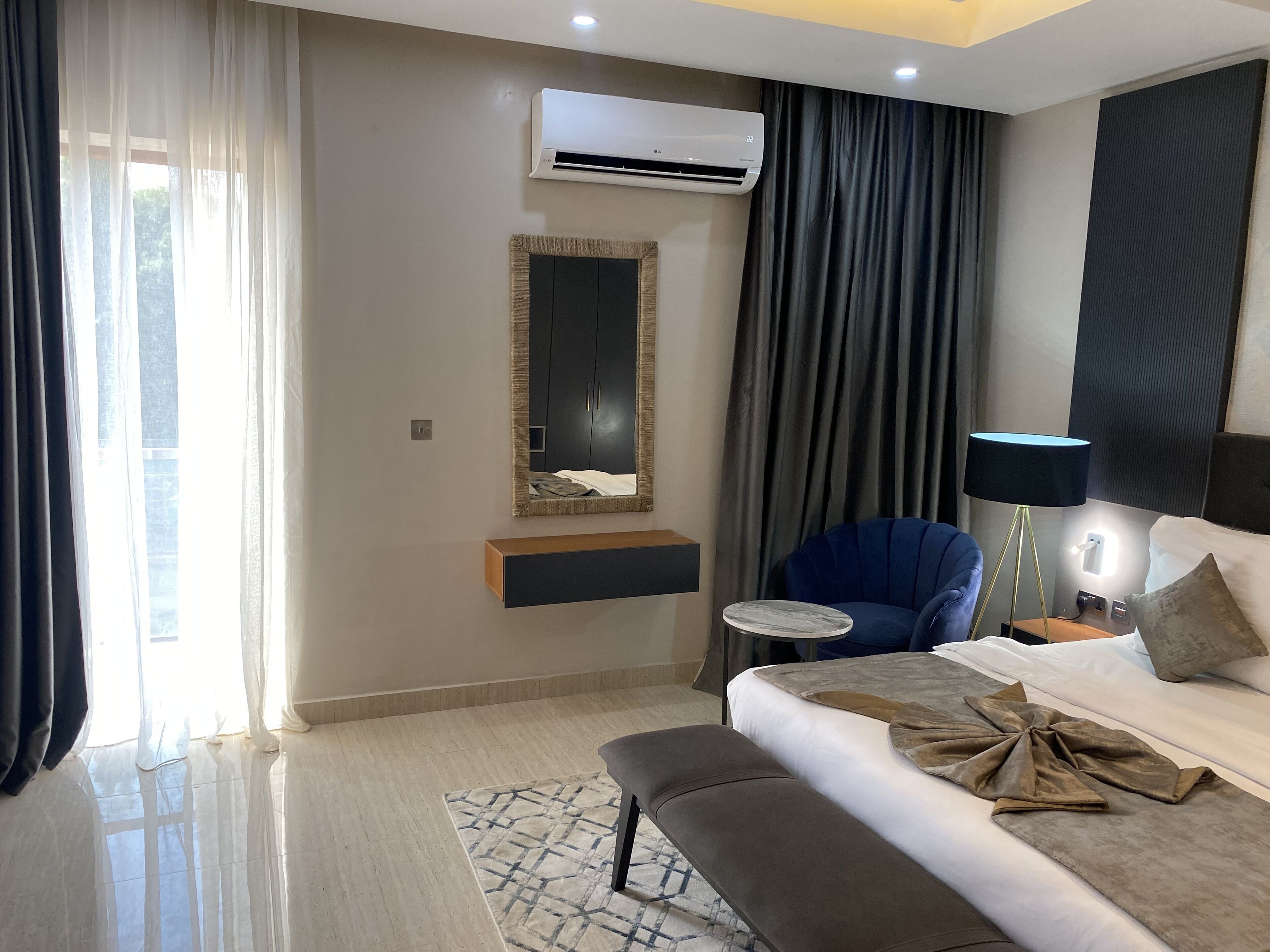 A modern hotel room with a large bed, a bedside table, a chair, an air conditioning unit, and a window with curtains.