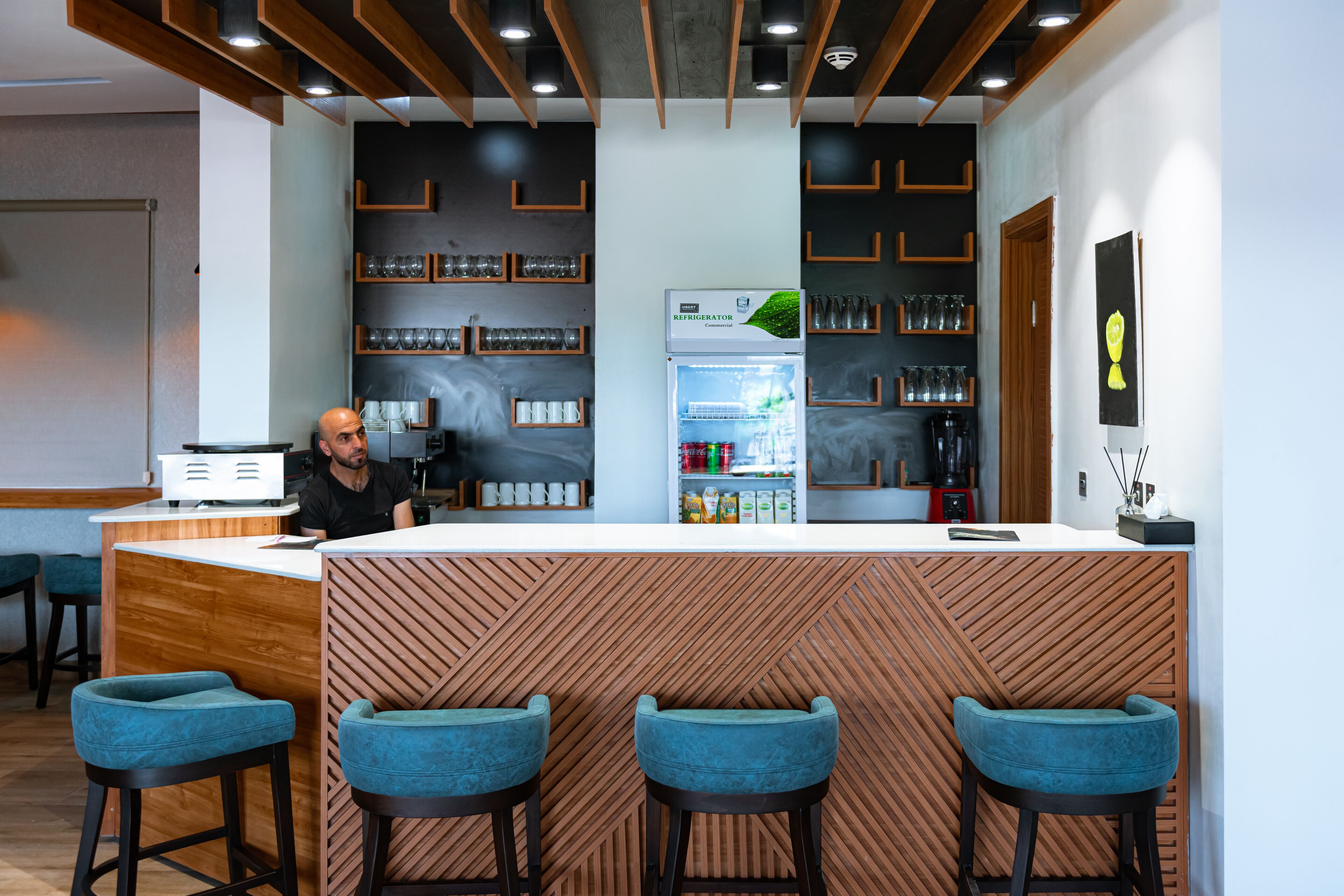 A modern bar area with a wooden counter, blue bar stools, and shelves with glasses and bottles.