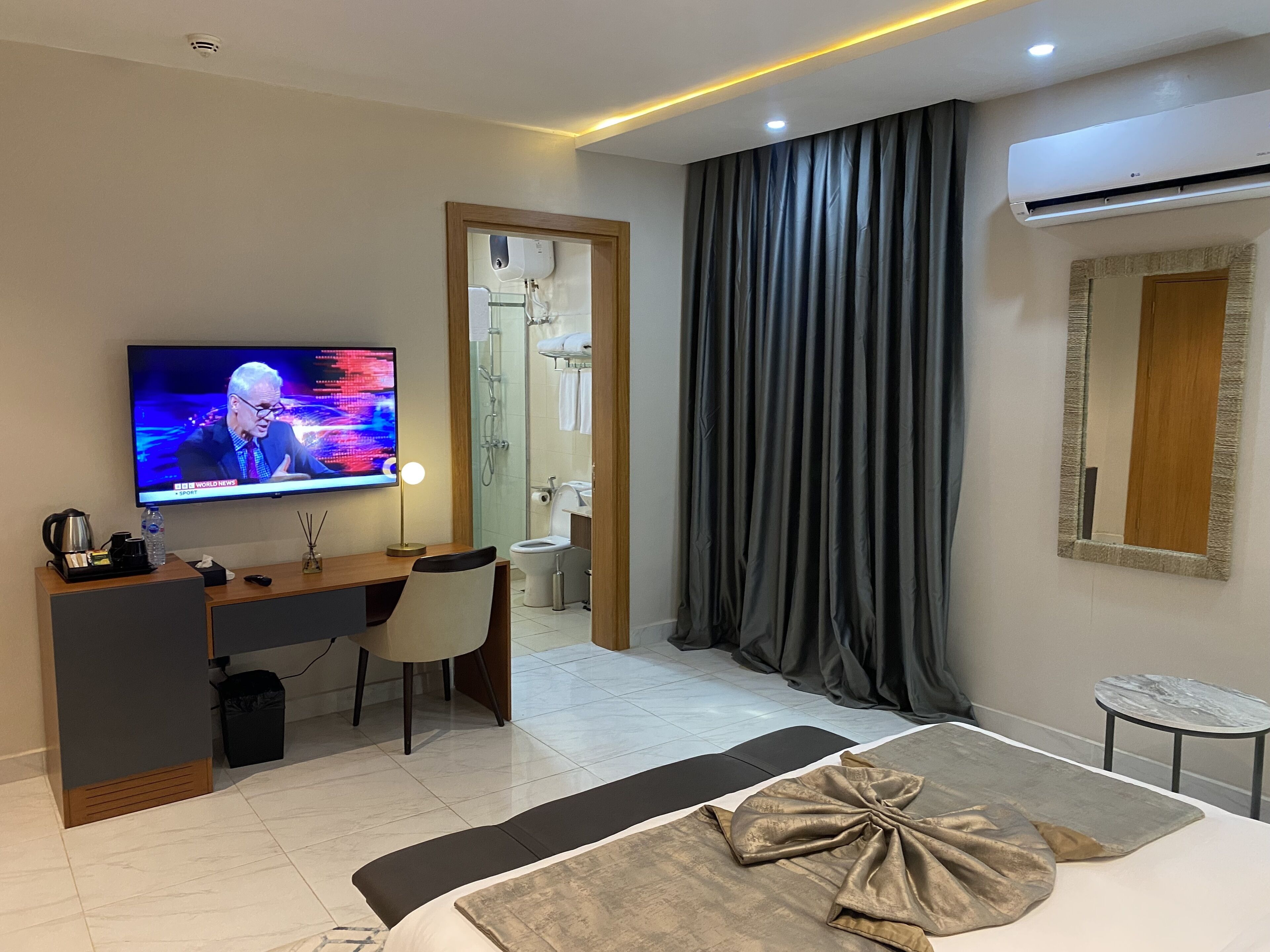 A modern hotel room with a flat-screen TV, a desk, a chair, a bed with a decorative pillow, a small table, and an air conditioning unit.