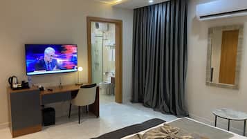 A modern hotel room with a flat-screen TV, a desk, a chair, a bed with a decorative pillow, a small table, and an air conditioning unit.