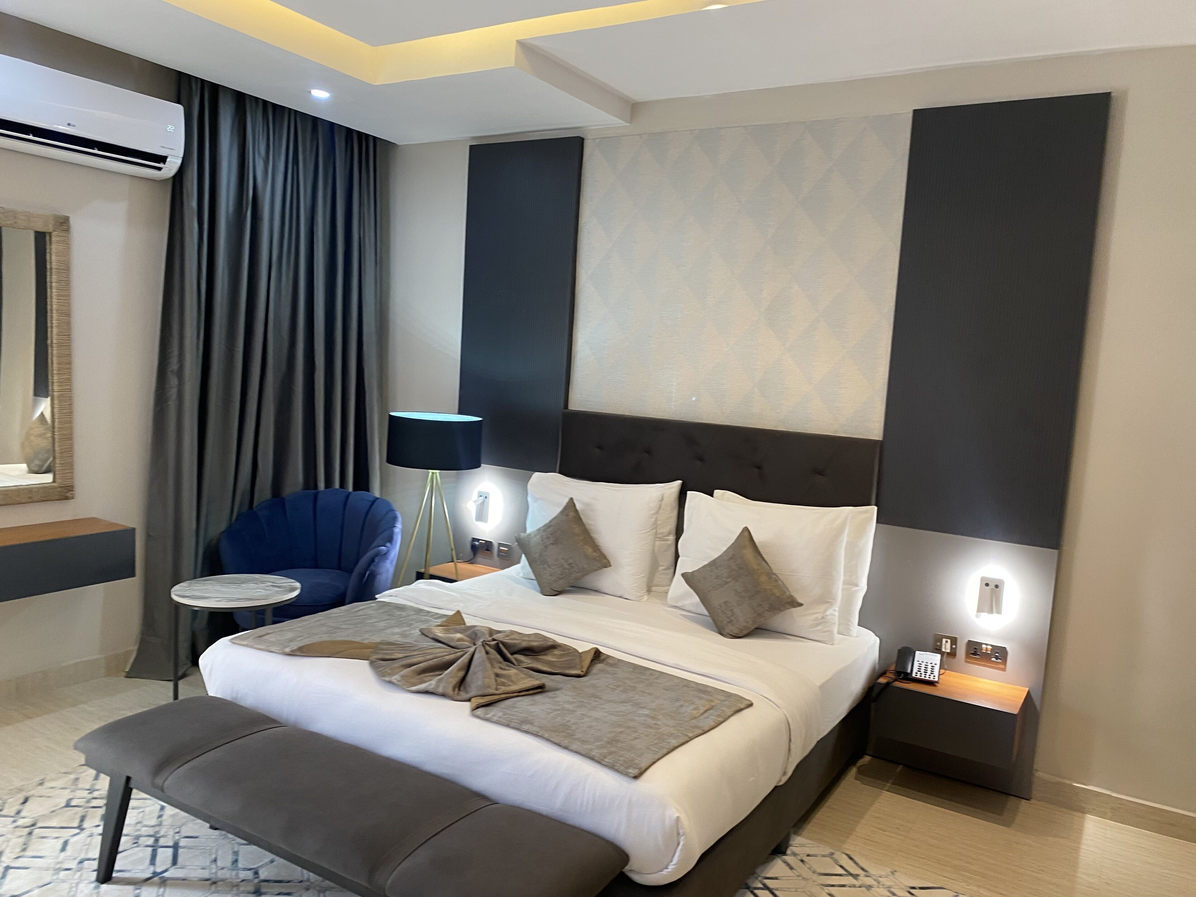 A modern hotel room with a large bed, a bedside table, a chair, an air conditioning unit, and a lamp.