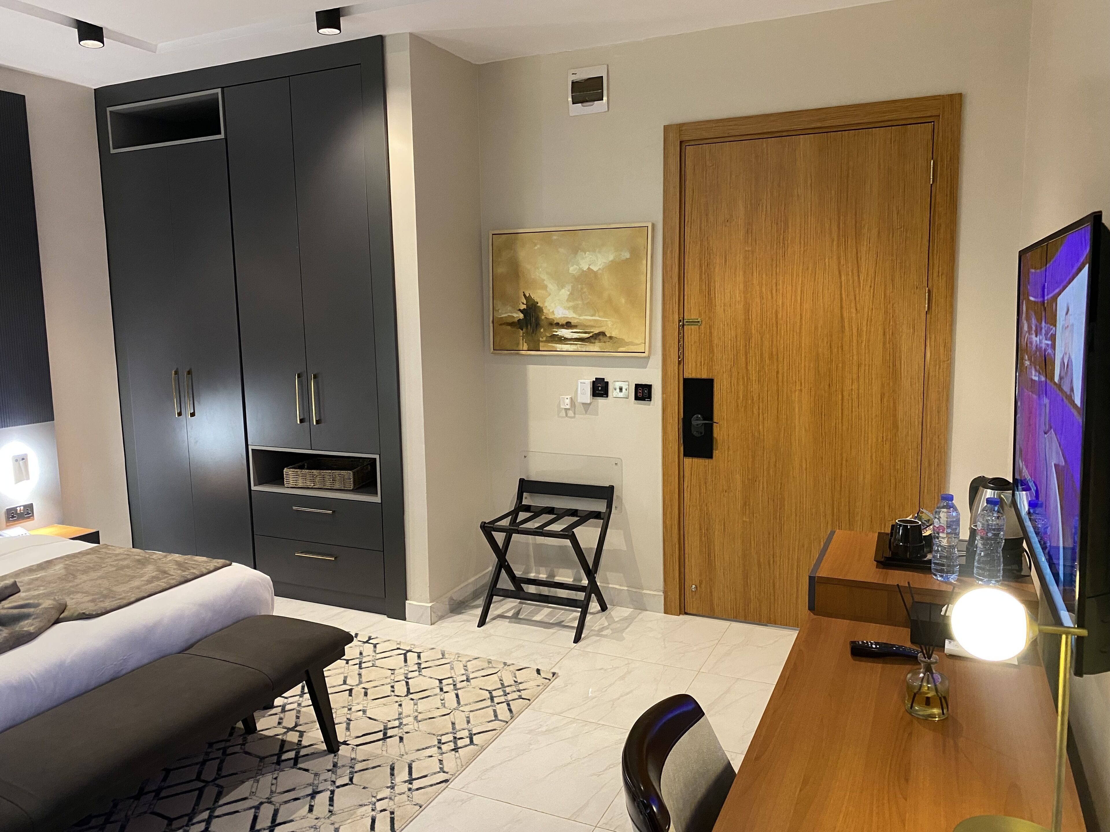 A modern hotel room with a bed, a desk with a lamp, a wooden door, and a painting on the wall.
