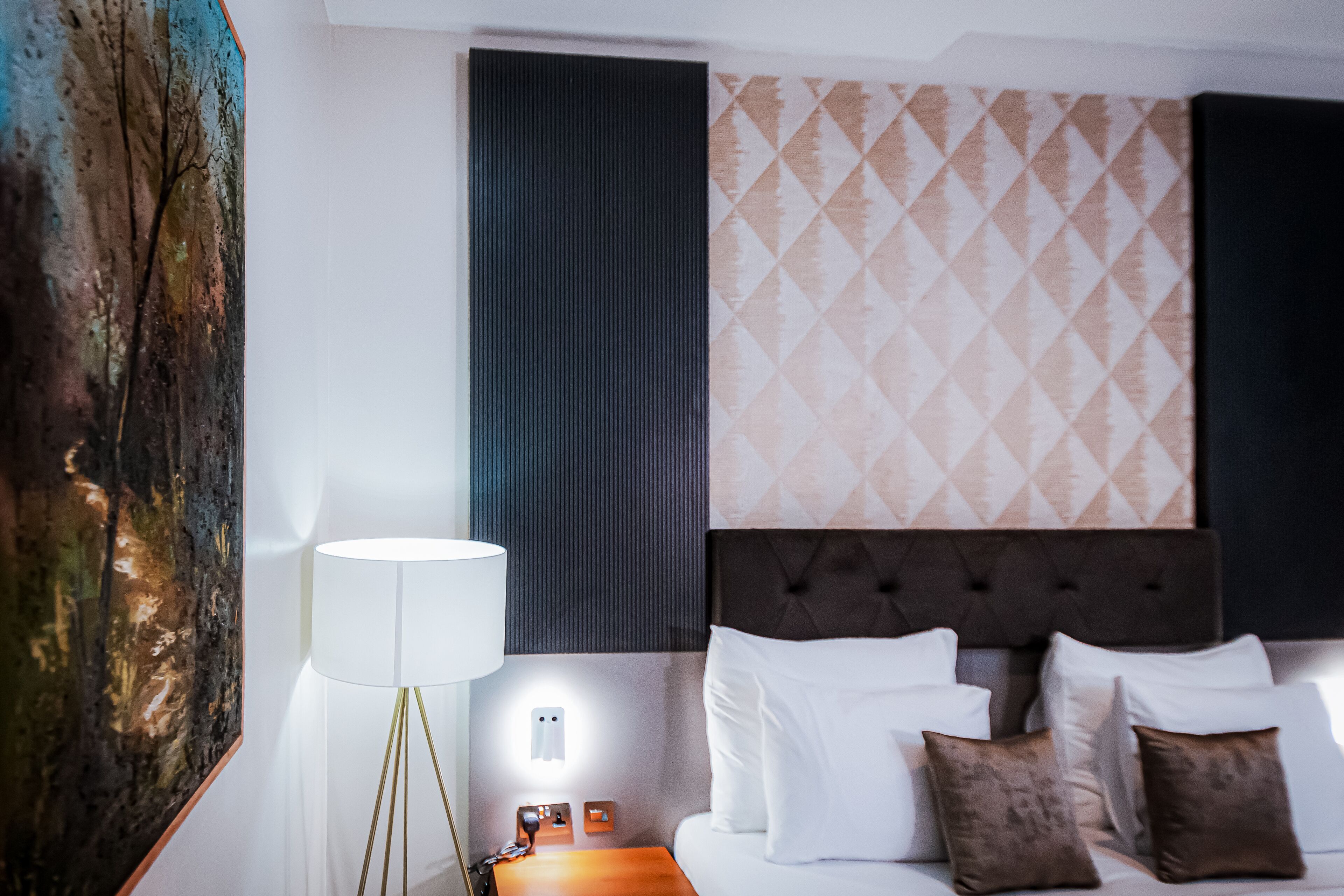 A hotel room with a bed, white pillows, a dark headboard, a wall with patterned wallpaper, and a floor lamp.