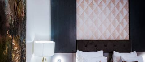 A hotel room with a bed, white pillows, a dark headboard, a wall with patterned wallpaper, and a floor lamp.