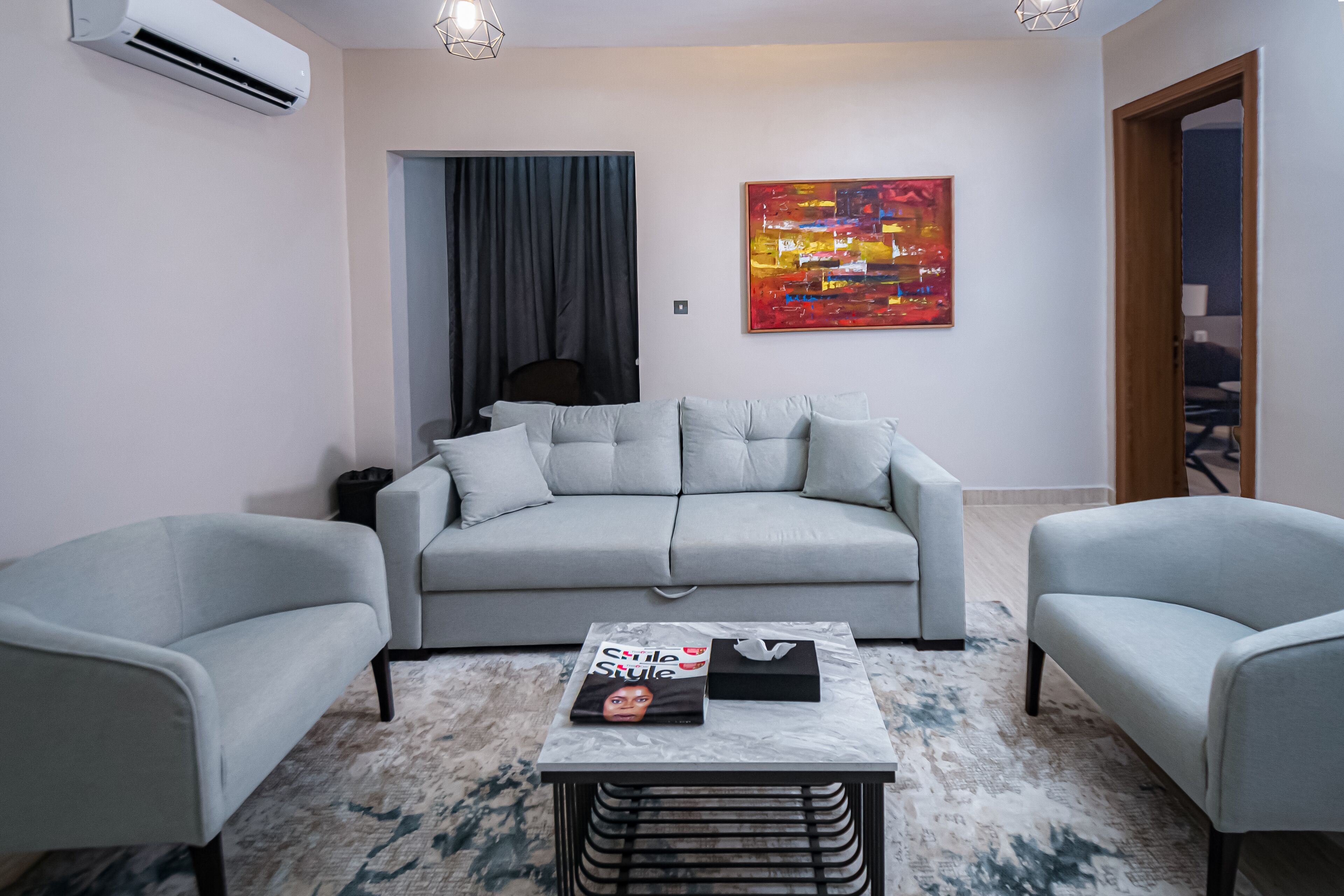 Una sala moderna con un sofá gris, dos sillones, una mesa de centro con una revista, y un cuadro abstracto y colorido en la pared.