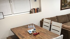 Un coin repas avec une table en bois, des chaises blanches et un banc. On peut voir des bouteilles d’eau sur la table.