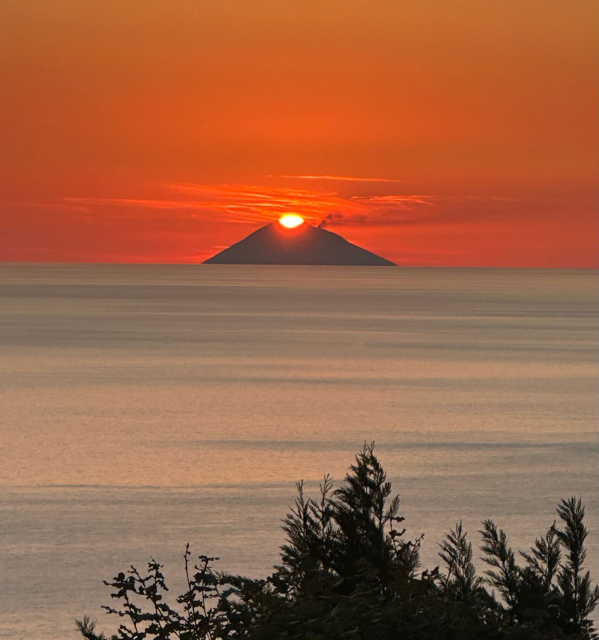A sunset over a calm sea with a silhouette of a mountain and some vegetation in the foreground.