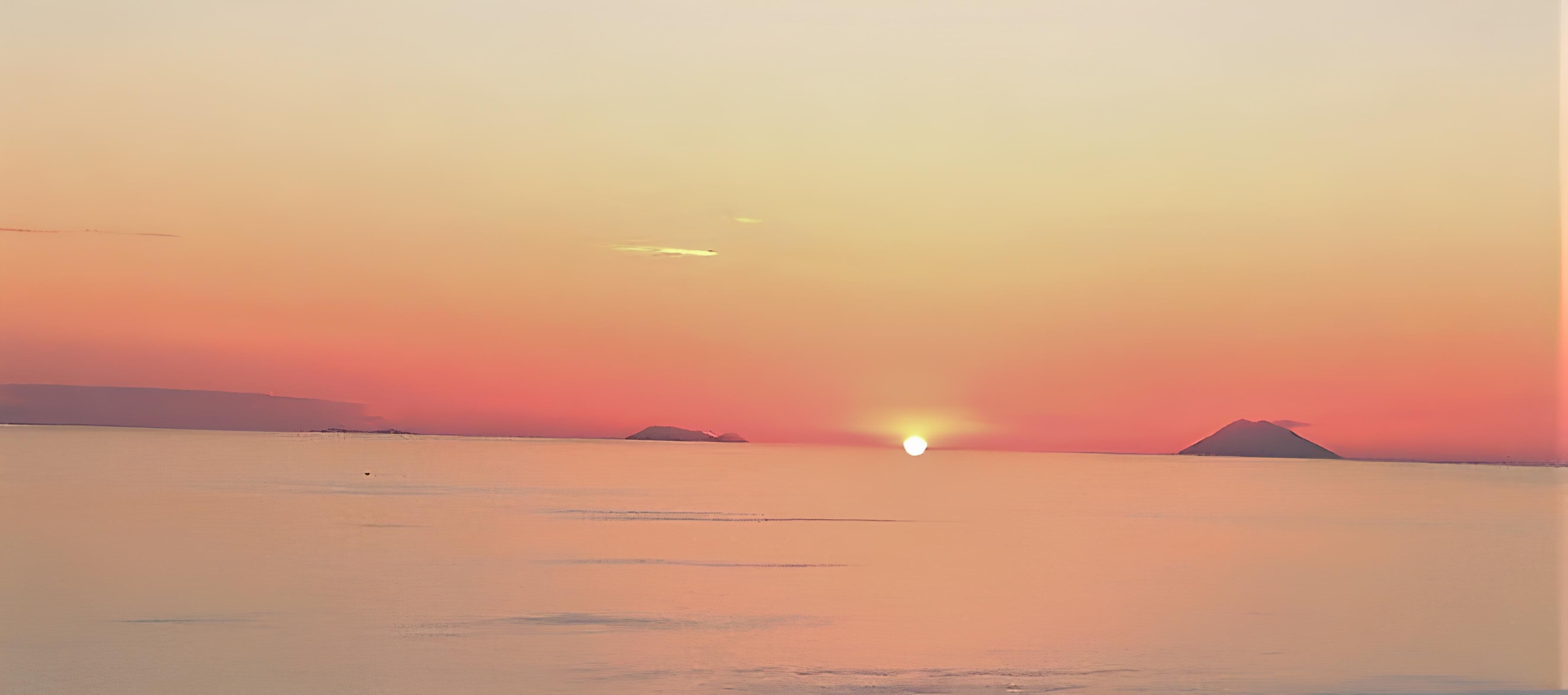 Un coucher de soleil sur une mer calme, avec des reliefs terrestres visibles au loin.