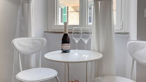 A white table with a bottle and two glasses, two white chairs, and a window with curtains. - APPARTAMENTO SANTI QUATTRO 1 - HOME AND MORE (Roma)