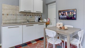 A modern kitchen with white cabinets, a tiled backsplash, and a wooden dining table with white chairs. A television displaying a show is mounted on the wall. - APPARTAMENTO SANTI QUATTRO 1 - HOME AND MORE (Roma)