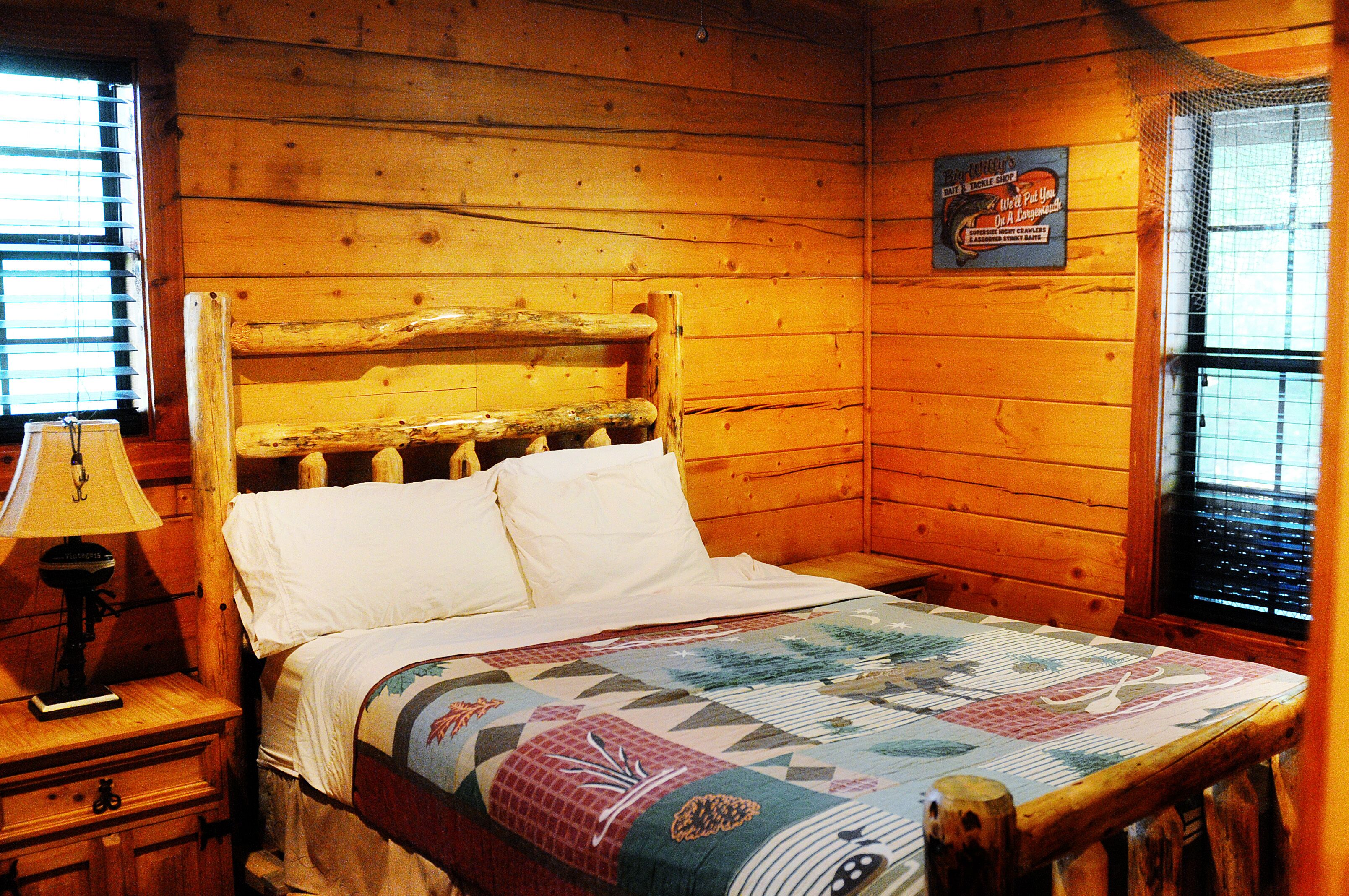 A wooden cabin room with a bed, a nightstand, a lamp, and a window with blinds.