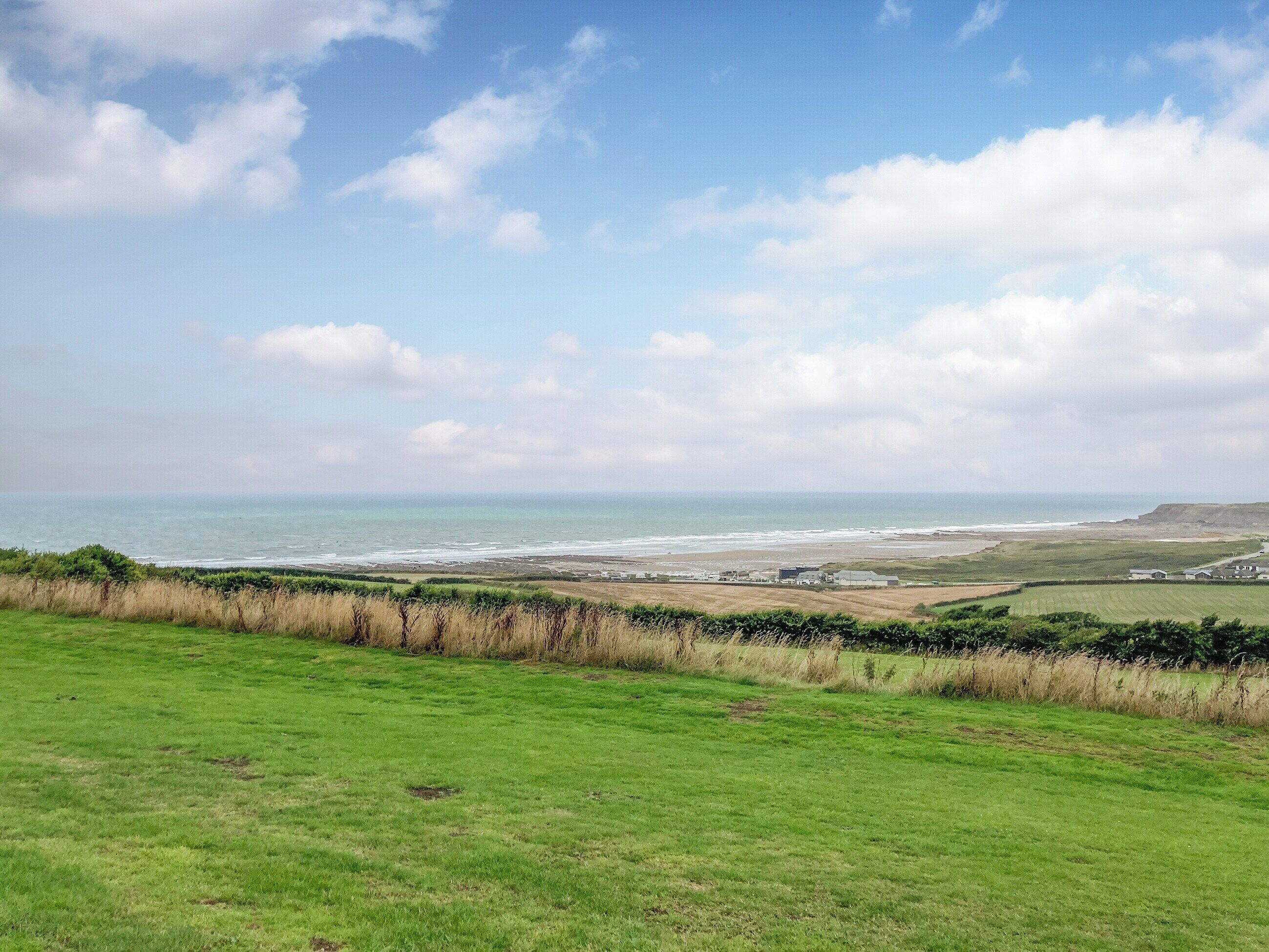Eine Küstenlandschaft mit einer grasbewachsenen Vordergrundebene, einem Strand und einer entfernten Meerblick unter einem teils bewölkten Himmel.
