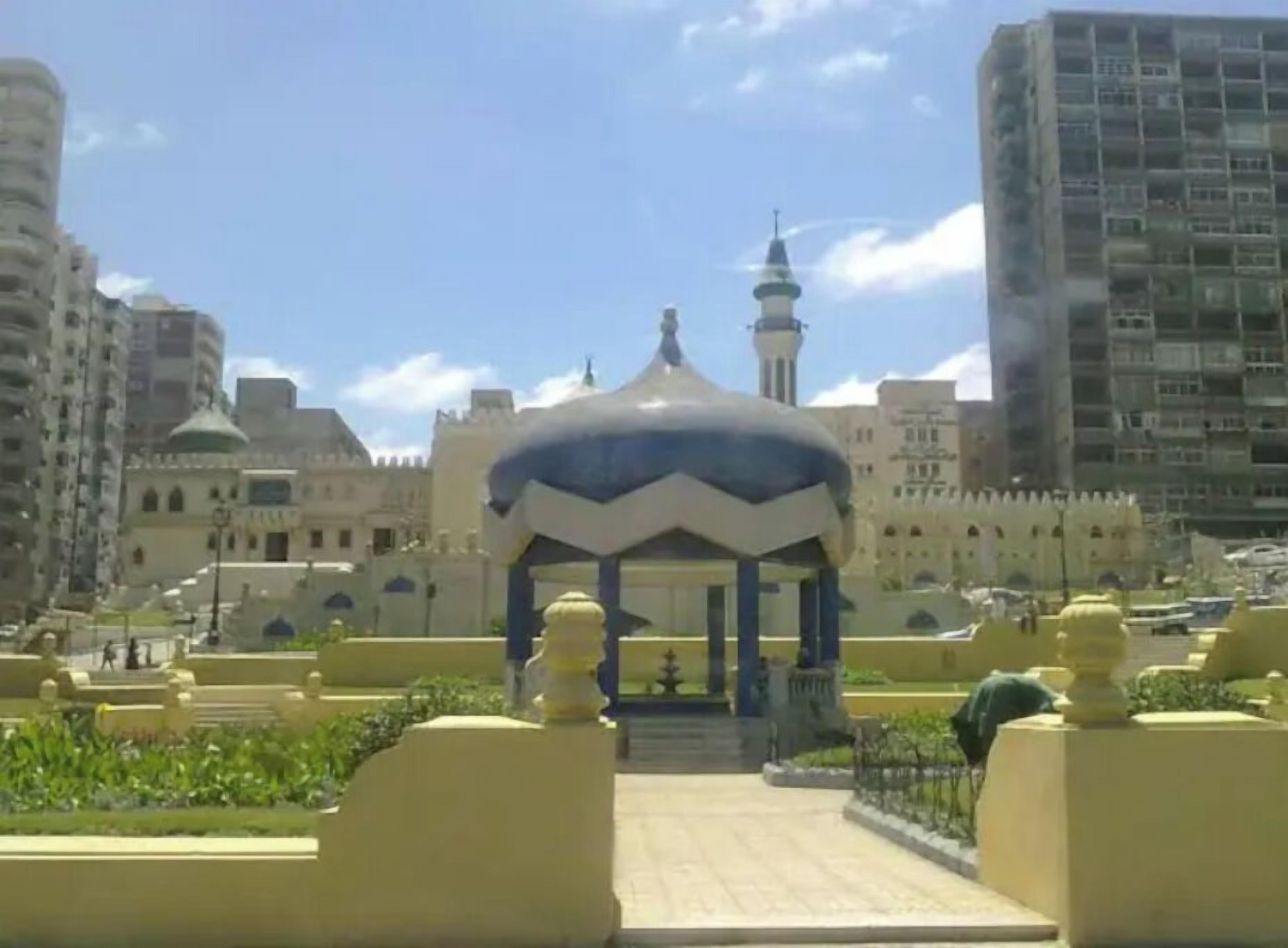 A building with a large central dome and a minaret, surrounded by greenery and modern high-rise buildings.