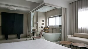 Une chambre d’hôtel moderne dotée d’un grand lit, d’un coin salon avec un fauteuil et de petites tables, d’une salle de bain fermée par des parois de verre et d’une grande fenêtre avec des rideaux.