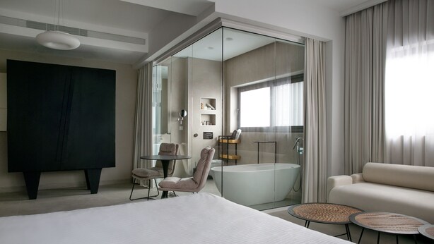 Ein modernes Hotelzimmer mit einem großen Bett, einer Sitzecke mit einem Sessel und kleinen Tischen, einem Glaskastenbad und einem großen Fenster mit Vorhängen.