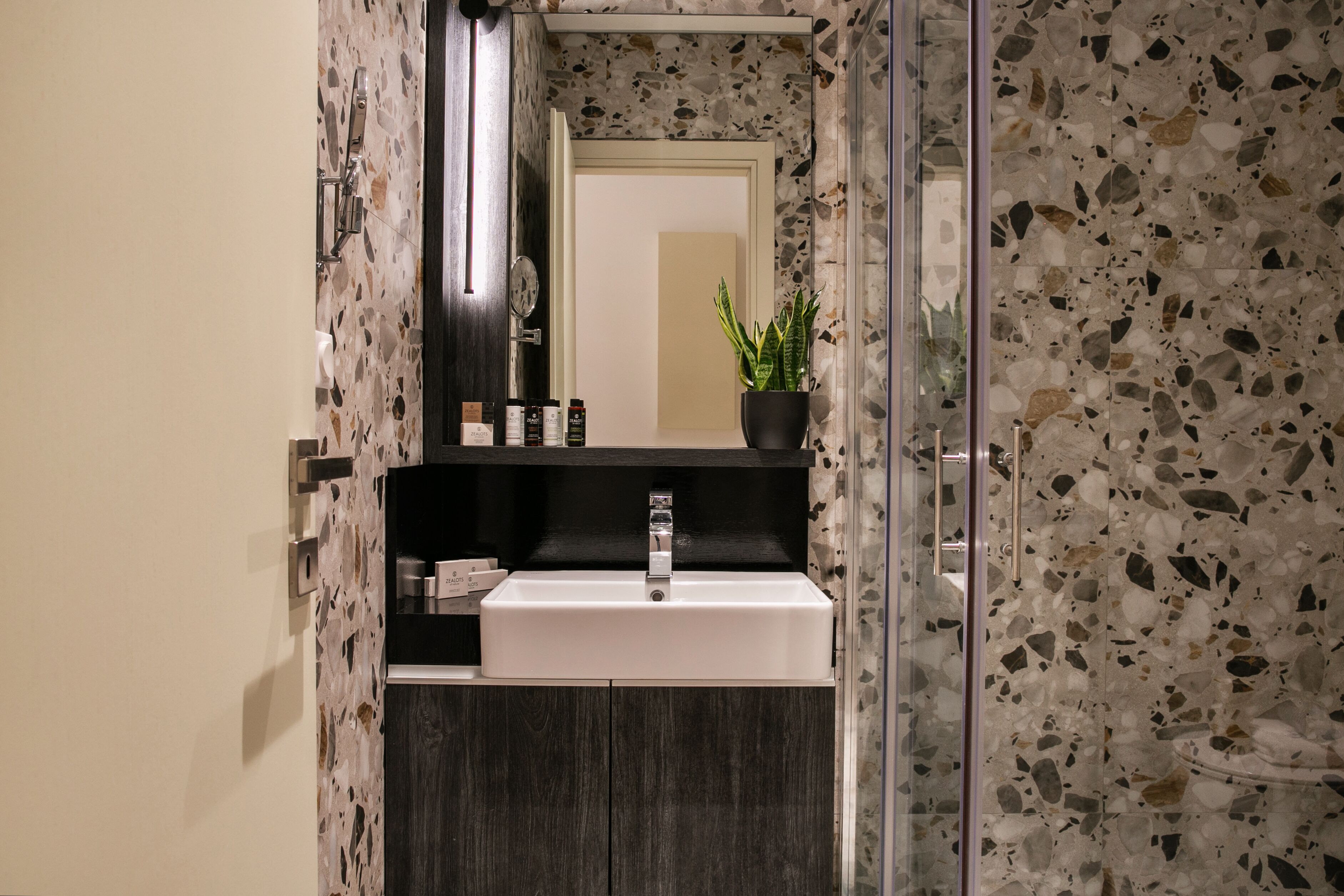 A modern bathroom with a rectangular sink, a mirror, a potted plant, and a shower area with a glass door.
