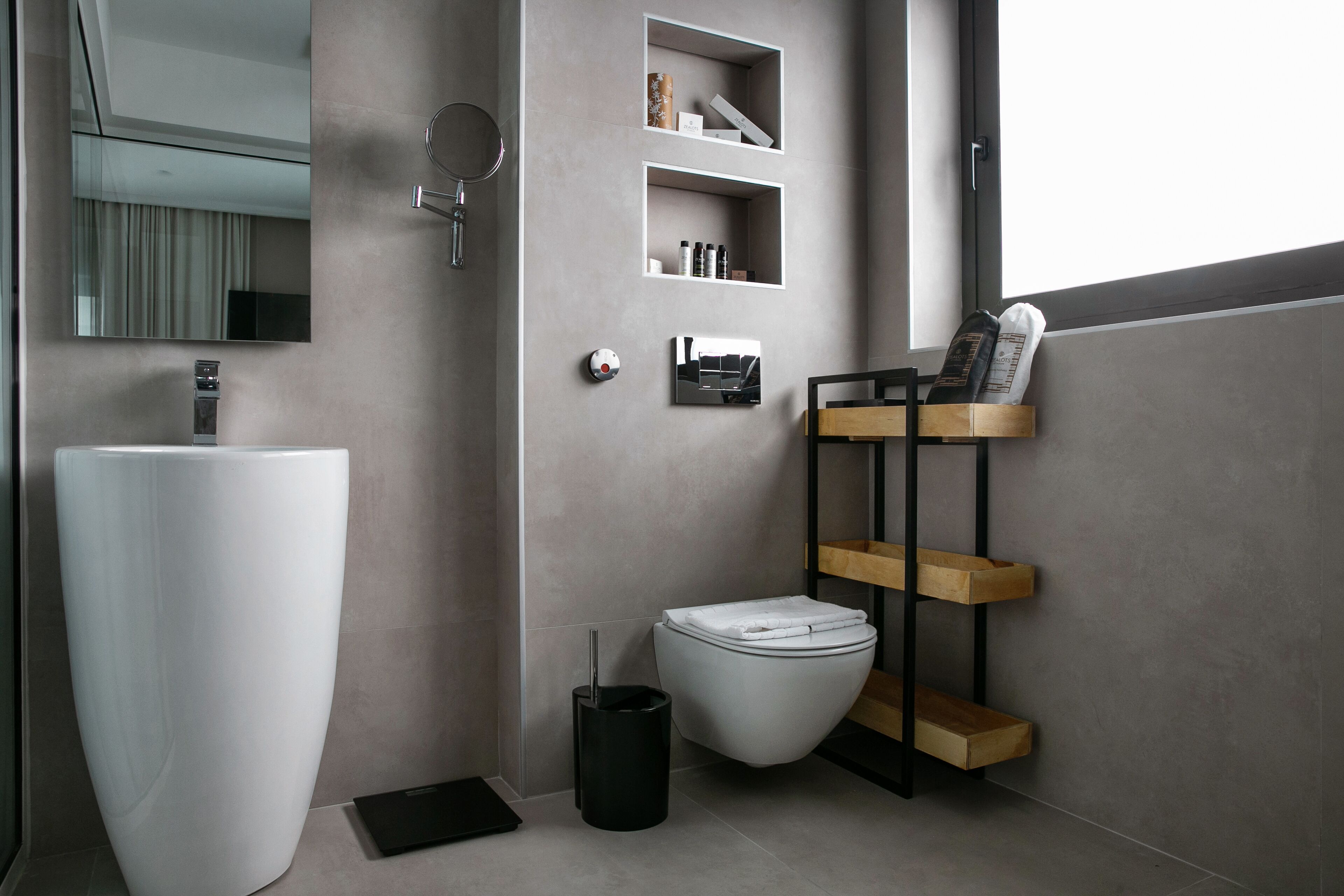 Ein modernes Badezimmer mit einem großen weißen Waschbecken, einer Toilette und einer schwarzen Mülltonne. Es gibt Regale mit Toilettenartikeln und einen Spiegel über dem Waschbecken.