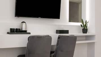 A modern room with a flat-screen TV mounted on the wall, a small table with a kettle and cups, two grey chairs, a potted plant, and a trash bin.