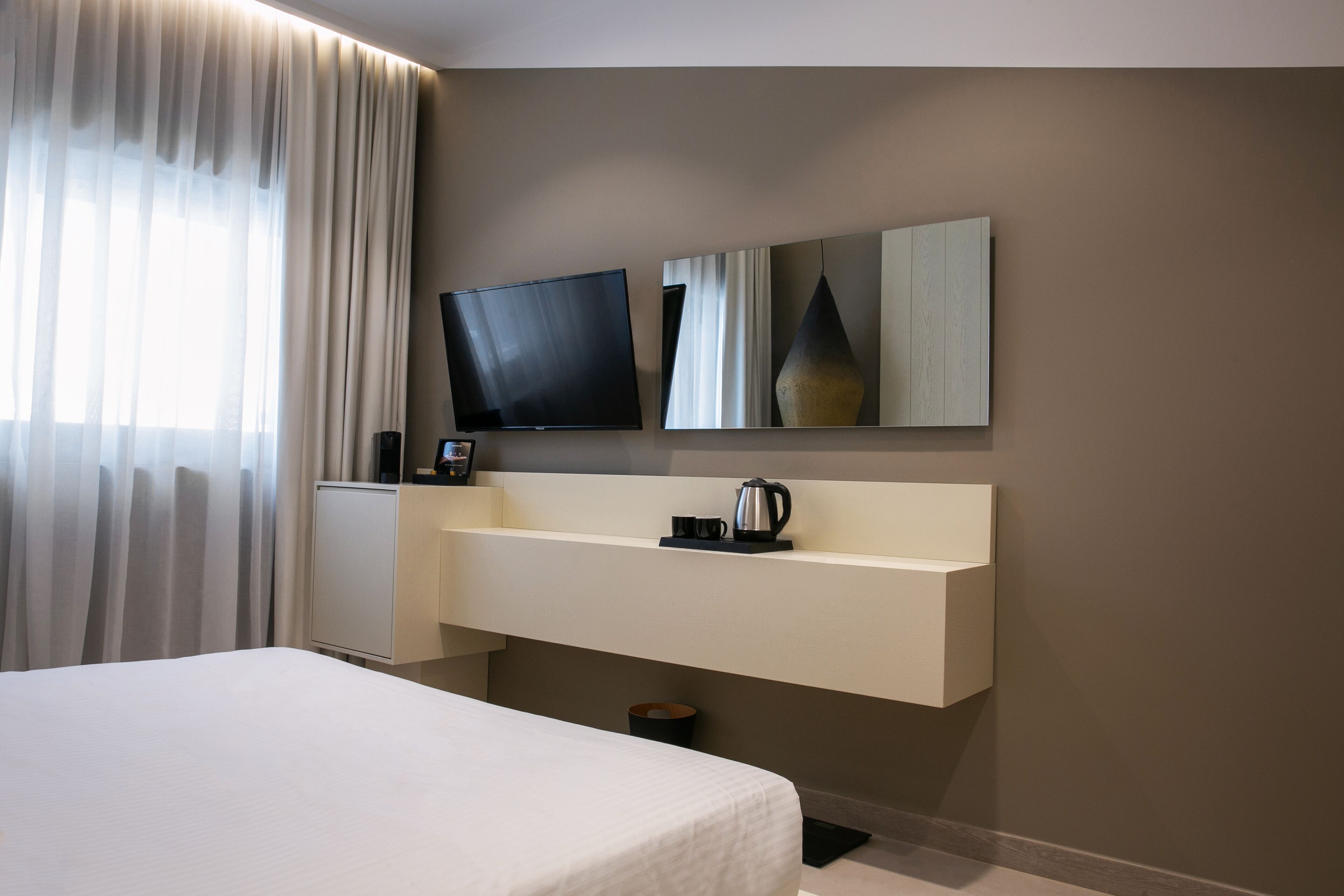 Una habitación de hotel moderna con una cama grande, un televisor de pantalla plana montado en la pared, una mesita de noche con un hervidor y un espejo grande sobre la mesa.