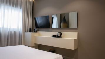 Una habitación de hotel moderna con una cama grande, un televisor de pantalla plana montado en la pared, una mesita de noche con un hervidor y un espejo grande sobre la mesa.