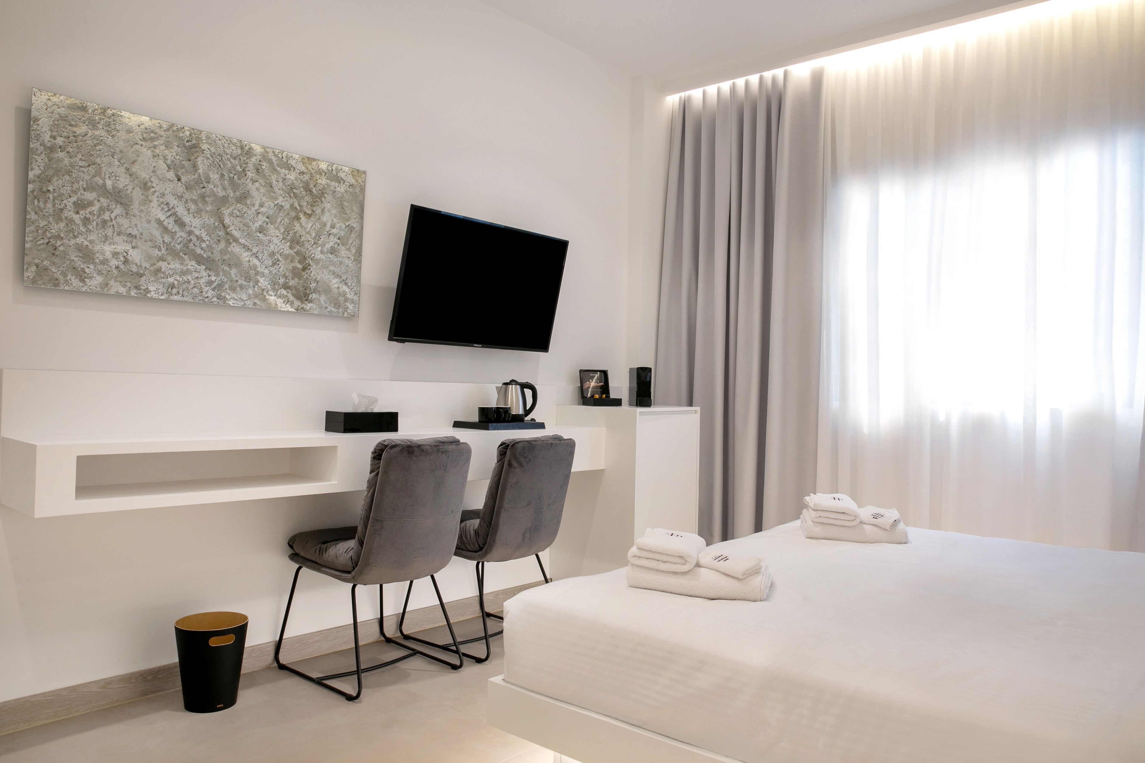 A modern hotel room with a flat-screen TV, a painting, a desk with two chairs, a bed with towels, and a large window with curtains.