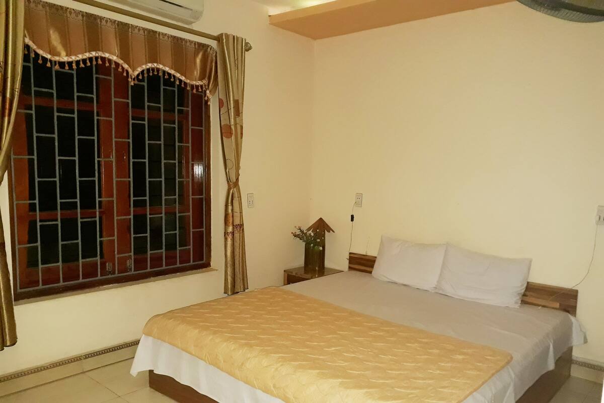 A bedroom with a bed, a window with curtains, a nightstand with a vase, and a wall socket.