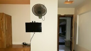 Een kamer met een houten bankje, een aan de muur bevestigde televisie en een staande ventilator.