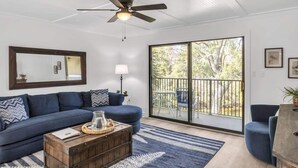 A living room with a blue sofa, a wooden coffee table, a ceiling fan, and a large sliding glass door leading to an outdoor balcony.