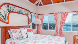 Una camera da letto con un letto di legno, biancheria da letto floreale, un'ampia finestra con tende e vista sul mare.