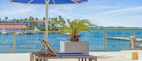 A sunlit outdoor lounge area with a blue umbrella, lounge chairs, and a palm plant.
