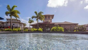 A swimming pool with clear water, surrounded by palm trees and a building with a tiled roof. - Pili Mai 7E - Top Floor 3 - Bed Suite On Golf Course - AC, Pool & Fitness Center (Koloa)