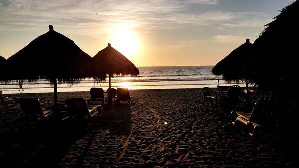 A beach with thatched umbrellas and lounge chairs, a sunset in the background, and the ocean.