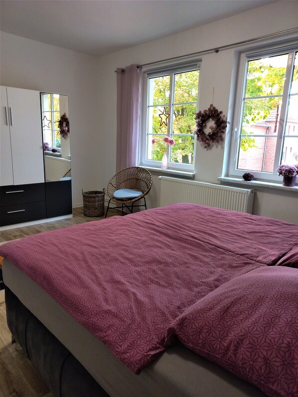 A bedroom with a bed, a chair, a wardrobe, and two windows with curtains.