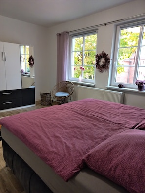 A bedroom with a bed, a chair, a wardrobe, and two windows with curtains. - Apartment 80m² cozy * stylish * with balcony (Wismar)