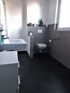 Een moderne badkamer met een wit bad, een toilet en een wastafel. Een potplant hangt aan de muur en een raam zorgt voor natuurlijk licht.