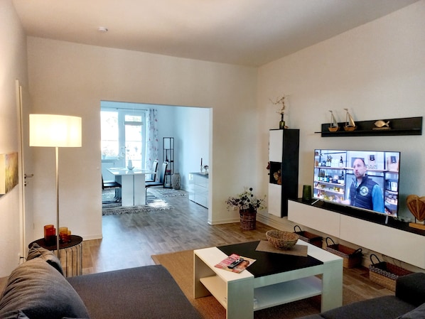 A modern living room with a flat-screen TV, a sofa, a coffee table, and a dining area in the background. - Apartment 80m² cozy * stylish * with balcony (Wismar)