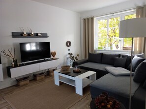 A modern living room with a flat-screen TV, a sofa, a coffee table, and a window with a view of trees.