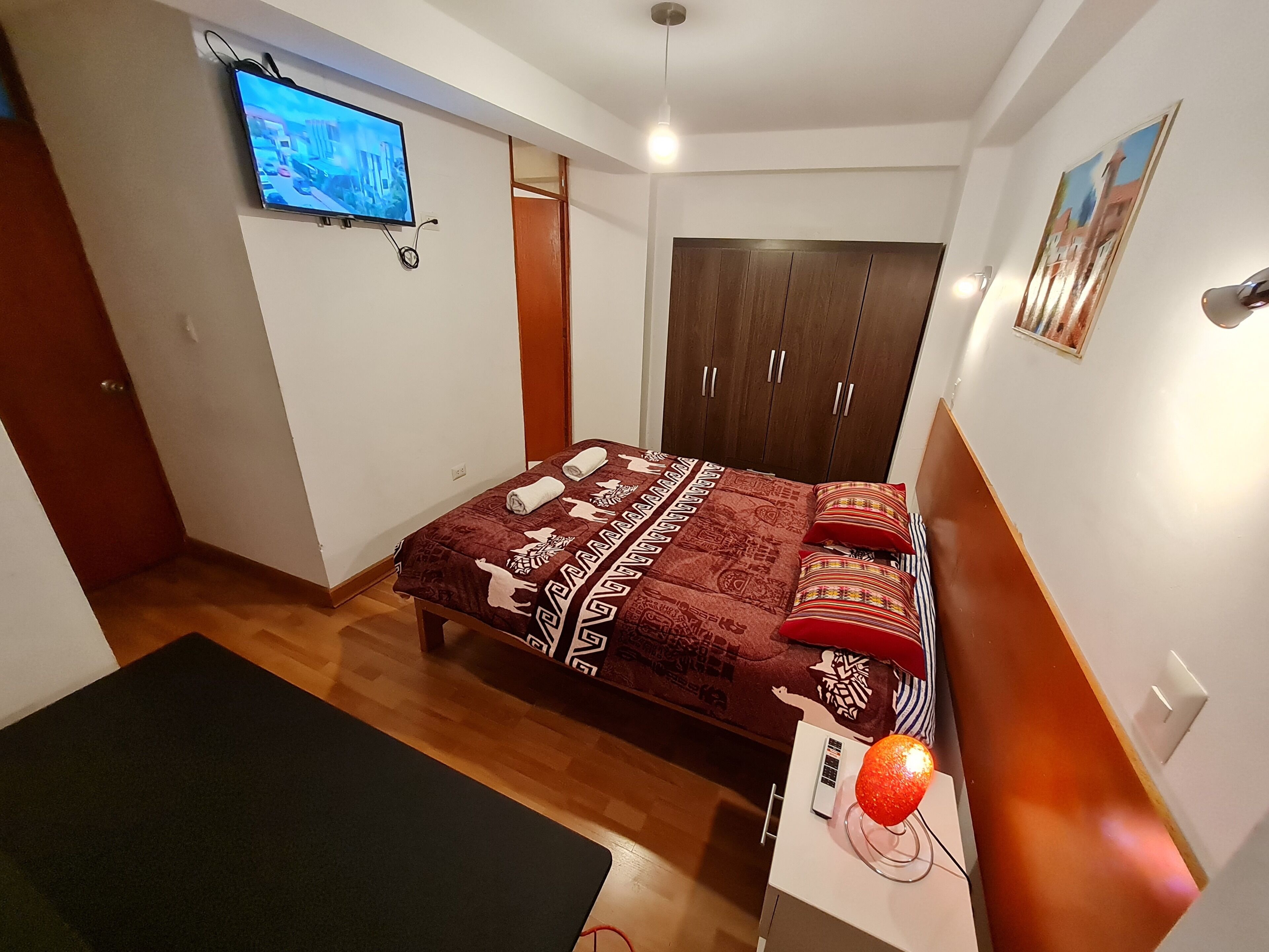 A bedroom with a bed, a TV mounted on the wall, a wardrobe, and a heater on a table.