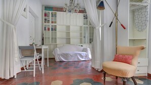 Ein Zimmer mit einem weißen Bett, einem Sessel, einem Schreibtisch und einem Bücherregal.