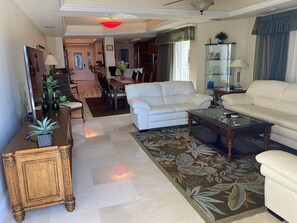 A spacious living room with a white sofa, a glass coffee table, and a dining area in the background.
