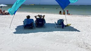 Una playa con un amplio toldo azul y blanco que proporciona sombra. Hay sillas de playa y una persona relajándose debajo del toldo.
