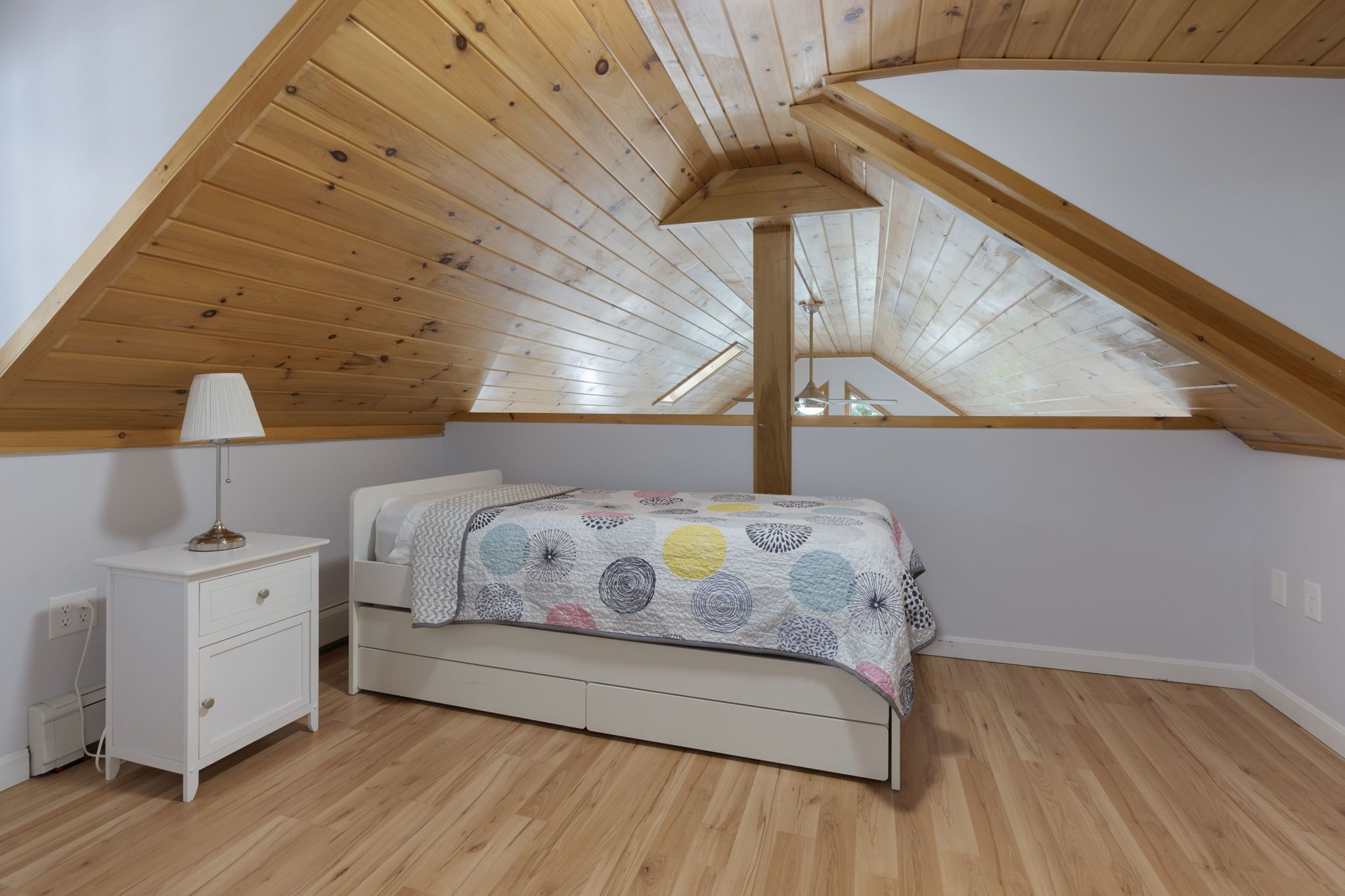 A bedroom with a sloped wooden ceiling, a bed with a patterned bedspread, a bedside table with a lamp, and a small white cabinet.