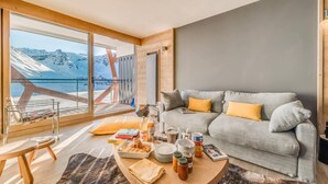 A modern living room with a grey sofa, yellow cushions, a coffee table with breakfast items, and a large window overlooking a snowy mountain. - 200m from ski slopes, 5th floor, view mountain, balcony, ski locker, 100m², Tignes (Tignes)